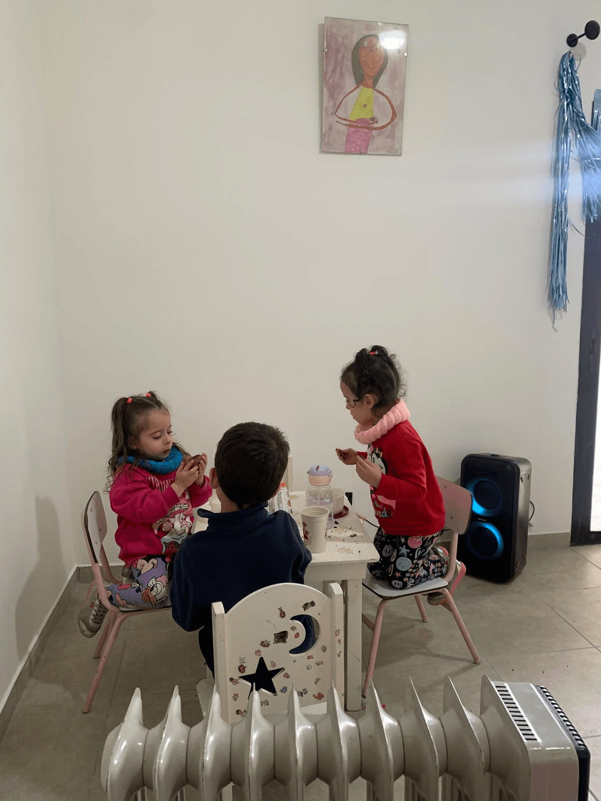 Tres niños merendando juntos en una mesita, con un dibujo colgado en la pared.